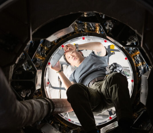NASA’s Unique Approach to Space Operations We look upward at an Asian man holding a silver tablet. He is wearing a gray t-shirt and green pants with a belt. He is halfway through a round opening. The inside of the craft he