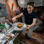 NASA Crew Marks Historic Achievement on Space Station Anne McClain, a white woman with short brown hair, floats while pointing downward with her right hand. Her left hand holds something that looks almost like a cake tin in place. There is a hamburger-shaped cake in the