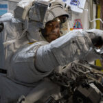 NASA astronauts don smiles while wearing spacesuits during mission. Chris Williams, an astronaut, is wearing a white spacesuit and helmet. He is facing the camera, but his body is turned to the right of the image. His arms are extended in front of him.