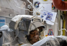 NASA astronauts don smiles while wearing spacesuits during mission. Chris Williams, an astronaut, is wearing a white spacesuit and helmet. He is facing the camera, but his body is turned to the right of the image. His arms are extended in front of him.