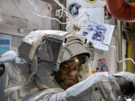 NASA astronauts don smiles while wearing spacesuits during mission. Chris Williams, an astronaut, is wearing a white spacesuit and helmet. He is facing the camera, but his body is turned to the right of the image. His arms are extended in front of him.