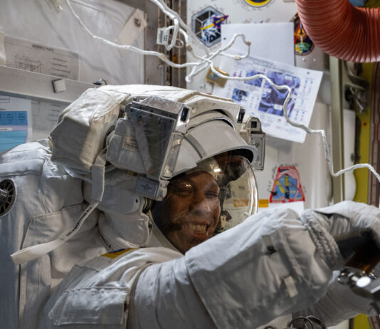 NASA astronauts don smiles while wearing spacesuits during mission. Chris Williams, an astronaut, is wearing a white spacesuit and helmet. He is facing the camera, but his body is turned to the right of the image. His arms are extended in front of him.