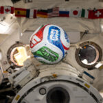 NASA Explores Intersection of Soccer and Space Science A white, red, blue, and green soccer ball floats inside the International Space Center. The FIFA logo is visible in the blue part of the ball facing the camera. The area in the background is mostly white, with a banner of country flags at the top of the photo.