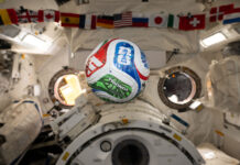 NASA Explores Intersection of Soccer and Space Science A white, red, blue, and green soccer ball floats inside the International Space Center. The FIFA logo is visible in the blue part of the ball facing the camera. The area in the background is mostly white, with a banner of country flags at the top of the photo.