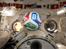 NASA Explores Intersection of Soccer and Space Science A white, red, blue, and green soccer ball floats inside the International Space Center. The FIFA logo is visible in the blue part of the ball facing the camera. The area in the background is mostly white, with a banner of country flags at the top of the photo.