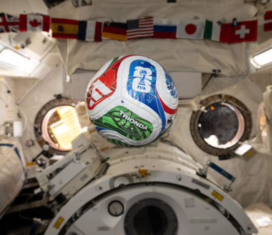 NASA Explores Intersection of Soccer and Space Science A white, red, blue, and green soccer ball floats inside the International Space Center. The FIFA logo is visible in the blue part of the ball facing the camera. The area in the background is mostly white, with a banner of country flags at the top of the photo.