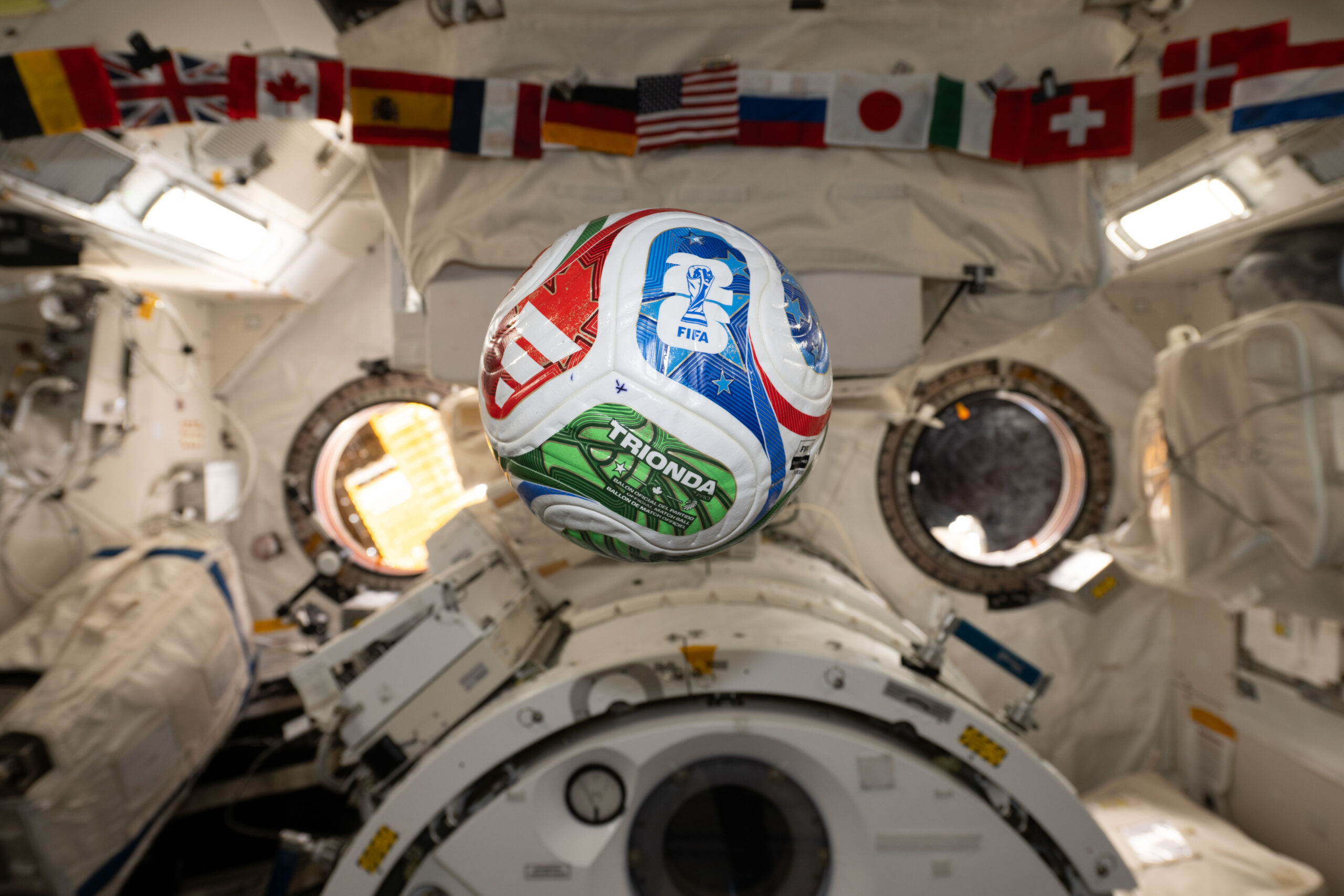 iss074e0349331.jpg A white, red, blue, and green soccer ball floats inside the International Space Center. The FIFA logo is visible in the blue part of the ball facing the camera. The area in the background is mostly white, with a banner of country flags at the top of the photo.