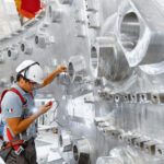 AI Advances Nuclear Fusion for Carbon-Free Energy Solutions A construction worker in a hard hat regarding a measuring device in his hand with a background of stainless steel wall with openings.