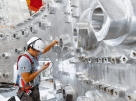 AI Advances Nuclear Fusion for Carbon-Free Energy Solutions A construction worker in a hard hat regarding a measuring device in his hand with a background of stainless steel wall with openings.