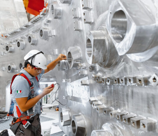 AI Advances Nuclear Fusion for Carbon-Free Energy Solutions A construction worker in a hard hat regarding a measuring device in his hand with a background of stainless steel wall with openings.