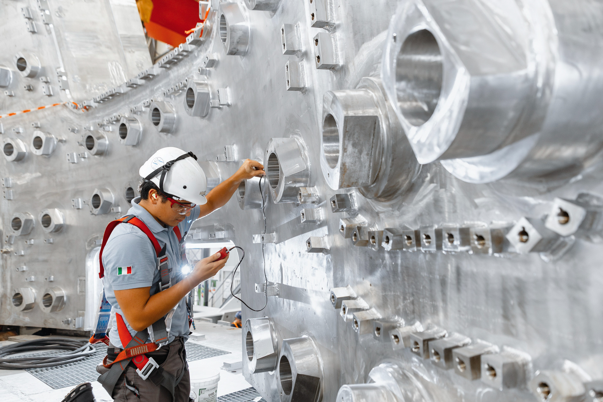 iter 13.jpg A construction worker in a hard hat regarding a measuring device in his hand with a background of stainless steel wall with openings.