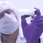 NASA’s Bennu asteroid sample contains life’s essential ingredients A scientist in a white coat, mask, hair net, and purple gloves holds up a clear vial with a black substance inside near the camera.
