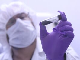 NASA’s Bennu asteroid sample contains life’s essential ingredients A scientist in a white coat, mask, hair net, and purple gloves holds up a clear vial with a black substance inside near the camera.