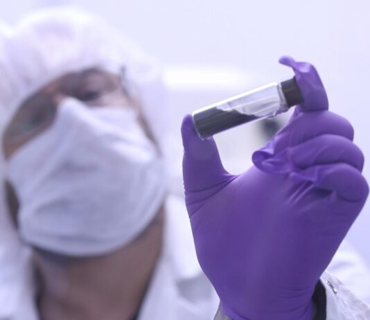 NASA’s Bennu asteroid sample contains life’s essential ingredients A scientist in a white coat, mask, hair net, and purple gloves holds up a clear vial with a black substance inside near the camera.