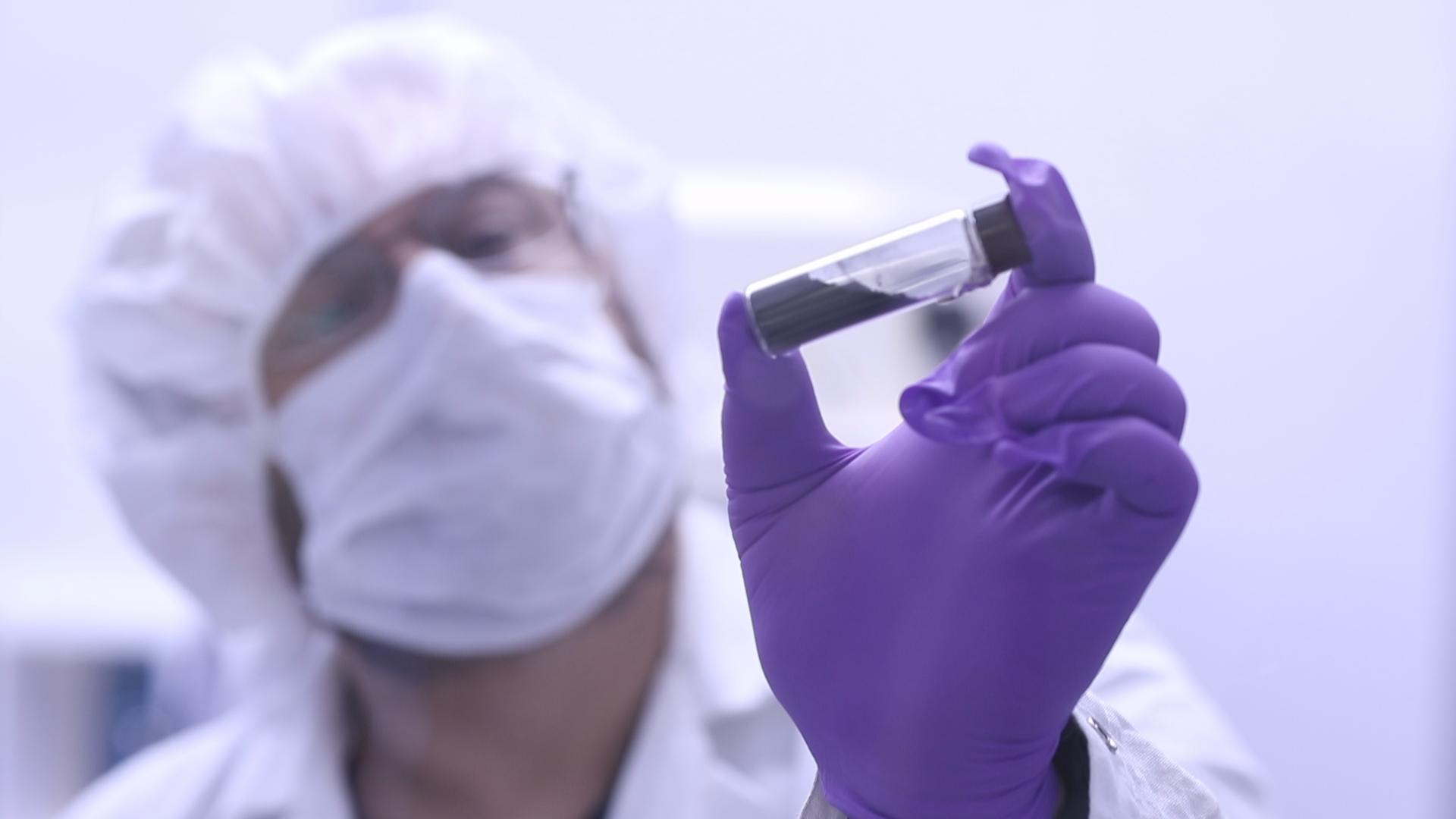 jason2large.jpg A scientist in a white coat, mask, hair net, and purple gloves holds up a clear vial with a black substance inside near the camera.