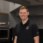 Joe Pavicic: NASA’s Artemis Mission Highlight Image shows man smiling in black shirt with NASA