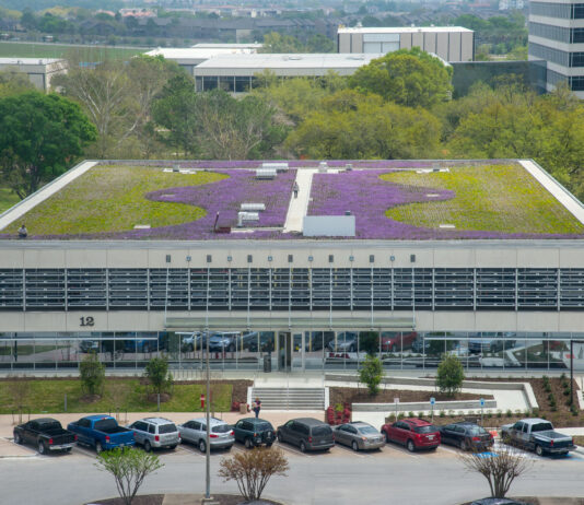 NASA’s Building 12 Hosts Thriving Pocket Prairie for Sustainability Aerial view of a building with a large rooftop garden featuring a mix of green and purple vegetation. The garden is designed in a curving, artistic pattern. In the background, other buildings and lush greenery are visible.