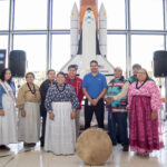 Kenneth Attocknie Connects Cultures at NASA Beyond Gaming A group of Indigenous people and a NASA employee pose together in front of a model of the Space Shuttle Endeavour. The individuals wear traditional clothing, including sashes and beaded garments.