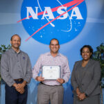 Jason Phillips’ Surprising Journey to NASA’s Johnson Procurement Jason Phillips stands between two managers on a stage with the NASA meatball in the background, holding an award.