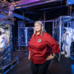 Caregivers at Johnson Balance Dual Roles with Community Support A woman stands inside a test chamber with several spacesuits.