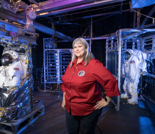 Caregivers at Johnson Balance Dual Roles with Community Support A woman stands inside a test chamber with several spacesuits.