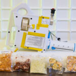 Artemis IV: NASA Develops Gateway Tech for Deep Space Meals The image shows a compact, handheld water dispenser device placed on a table, with several vacuum-sealed packets of food arranged in front of it. The device is white with yellow tape and has various labels and instructions on its surface. It includes a large, graduated syringe-like component marked in milliliters, used for precise water measurement. The food packets in front of the device contain different types of rehydratable meals in various colors and textures, indicating the preparation of food for consumption in a space environment. The background features a white grid pattern, enhancing the focus on the device and food packets. This device is being developed for astronauts who live and work aboard Gateway.