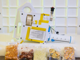 Artemis IV: NASA Develops Gateway Tech for Deep Space Meals The image shows a compact, handheld water dispenser device placed on a table, with several vacuum-sealed packets of food arranged in front of it. The device is white with yellow tape and has various labels and instructions on its surface. It includes a large, graduated syringe-like component marked in milliliters, used for precise water measurement. The food packets in front of the device contain different types of rehydratable meals in various colors and textures, indicating the preparation of food for consumption in a space environment. The background features a white grid pattern, enhancing the focus on the device and food packets. This device is being developed for astronauts who live and work aboard Gateway.