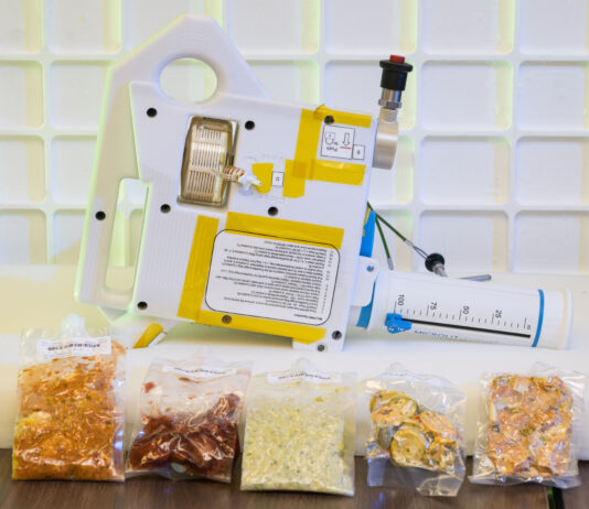 Artemis IV: NASA Develops Gateway Tech for Deep Space Meals The image shows a compact, handheld water dispenser device placed on a table, with several vacuum-sealed packets of food arranged in front of it. The device is white with yellow tape and has various labels and instructions on its surface. It includes a large, graduated syringe-like component marked in milliliters, used for precise water measurement. The food packets in front of the device contain different types of rehydratable meals in various colors and textures, indicating the preparation of food for consumption in a space environment. The background features a white grid pattern, enhancing the focus on the device and food packets. This device is being developed for astronauts who live and work aboard Gateway.