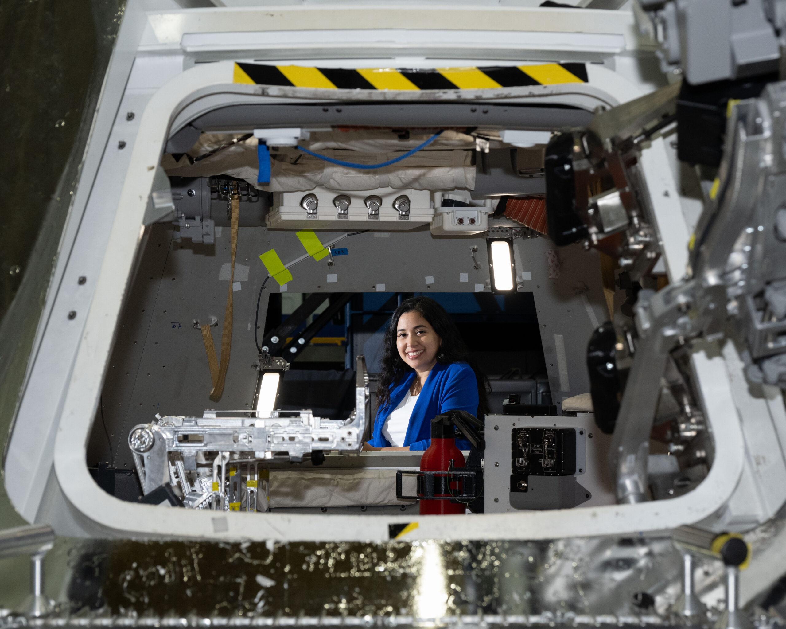 jsc2024e060575 alt.jpg Zaida Hernandez, a hispanic woman with long wavy brown hair and dark brown eyes, smiles brightly at the camera while sitting inside a mock up of NASA