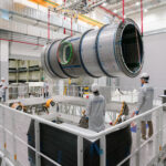 Lunar Module Heads to U.S. for NASA’s Artemis IV Mission A large cylindrical module is suspended by red straps as it is lowered onto a stand in a cleanroom at Thales Alenia Space in Turin, Italy. Engineers and technicians in white lab coats and helmets observe and guide the process from an elevated platform. The cleanroom features metal walkways and bright overhead lighting.