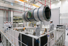 Lunar Module Heads to U.S. for NASA’s Artemis IV Mission A large cylindrical module is suspended by red straps as it is lowered onto a stand in a cleanroom at Thales Alenia Space in Turin, Italy. Engineers and technicians in white lab coats and helmets observe and guide the process from an elevated platform. The cleanroom features metal walkways and bright overhead lighting.
