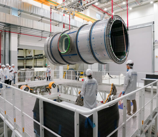 Lunar Module Heads to U.S. for NASA’s Artemis IV Mission A large cylindrical module is suspended by red straps as it is lowered onto a stand in a cleanroom at Thales Alenia Space in Turin, Italy. Engineers and technicians in white lab coats and helmets observe and guide the process from an elevated platform. The cleanroom features metal walkways and bright overhead lighting.