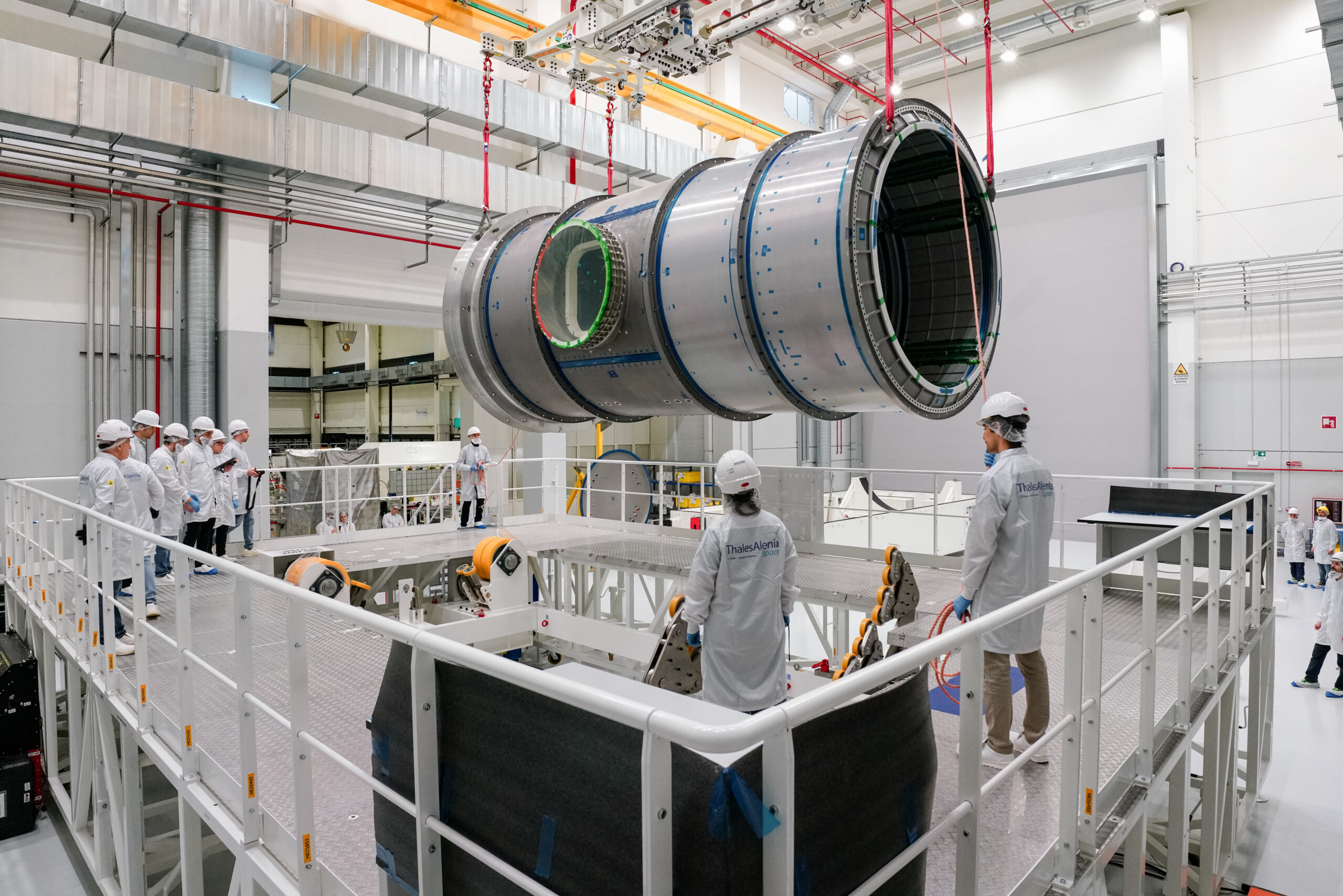jsc2025e003426.jpg A large cylindrical module is suspended by red straps as it is lowered onto a stand in a cleanroom at Thales Alenia Space in Turin, Italy. Engineers and technicians in white lab coats and helmets observe and guide the process from an elevated platform. The cleanroom features metal walkways and bright overhead lighting.