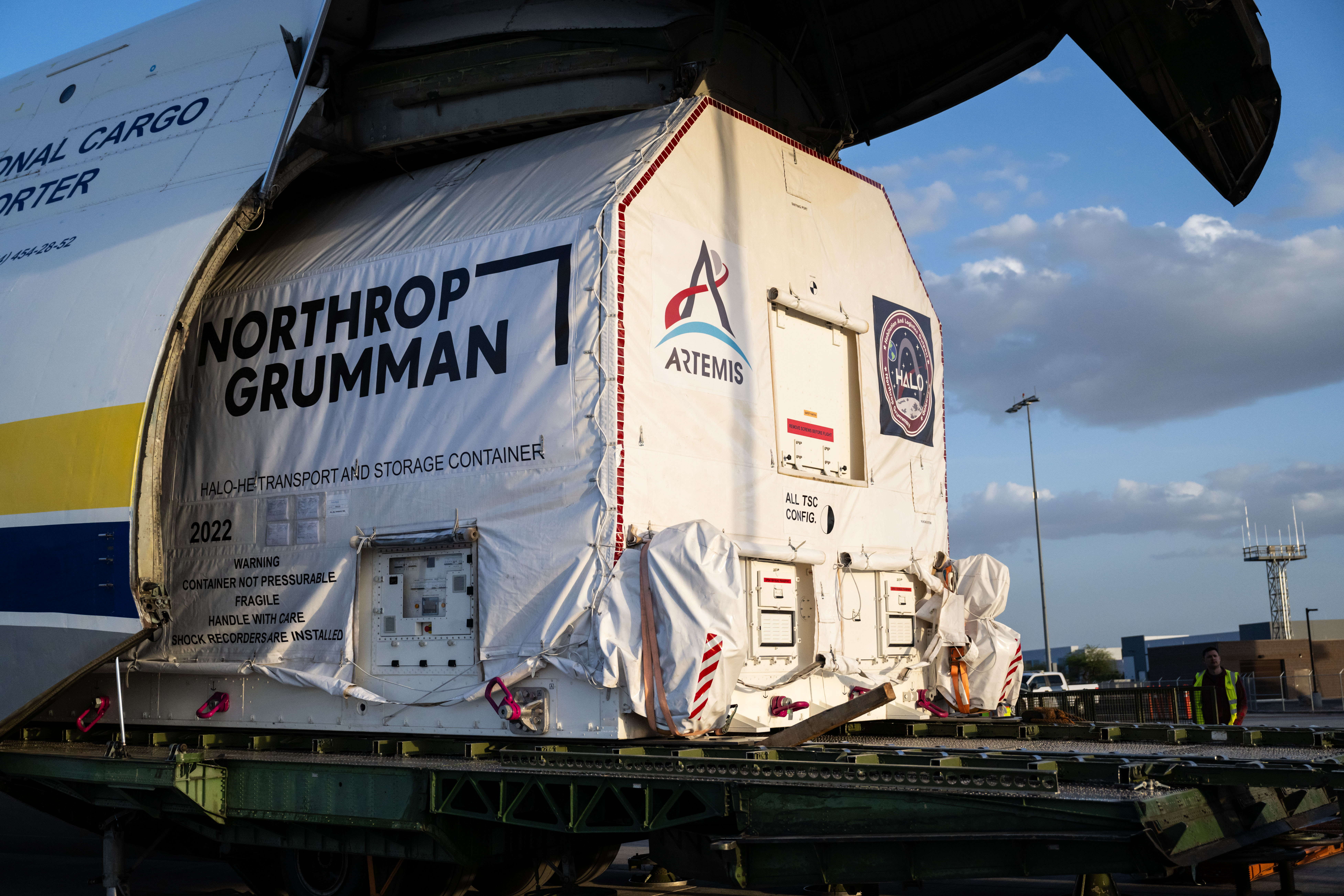 jsc2025e035259.jpg A large cargo aircraft with its nose lifted open reveals a wrapped container being offloaded. The container is marked with the logo for NASA