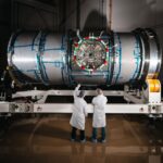 NASA’s Artemis Lunar Station Enters Final Testing Phase Two technicians in cleanroom attire stand in front of HALO (Habitation and Logistics Outpost) at Northrop Grumman’s facility in Gilbert, Arizona. The cylindrical metallic module, recently unboxed, rests on a white stand. One technician points toward the front of the module.