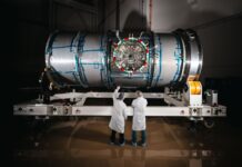 NASA’s Artemis Lunar Station Enters Final Testing Phase Two technicians in cleanroom attire stand in front of HALO (Habitation and Logistics Outpost) at Northrop Grumman’s facility in Gilbert, Arizona. The cylindrical metallic module, recently unboxed, rests on a white stand. One technician points toward the front of the module.