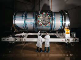 NASA’s Artemis Lunar Station Enters Final Testing Phase Two technicians in cleanroom attire stand in front of HALO (Habitation and Logistics Outpost) at Northrop Grumman’s facility in Gilbert, Arizona. The cylindrical metallic module, recently unboxed, rests on a white stand. One technician points toward the front of the module.