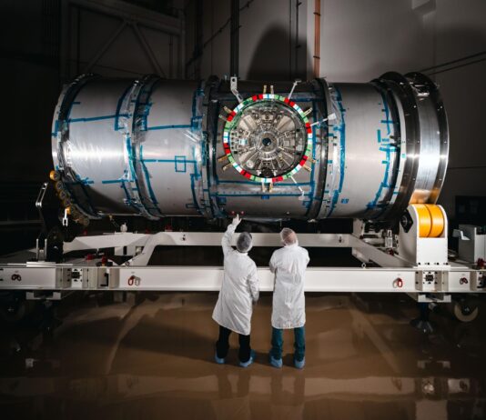 NASA’s Artemis Lunar Station Enters Final Testing Phase Two technicians in cleanroom attire stand in front of HALO (Habitation and Logistics Outpost) at Northrop Grumman’s facility in Gilbert, Arizona. The cylindrical metallic module, recently unboxed, rests on a white stand. One technician points toward the front of the module.