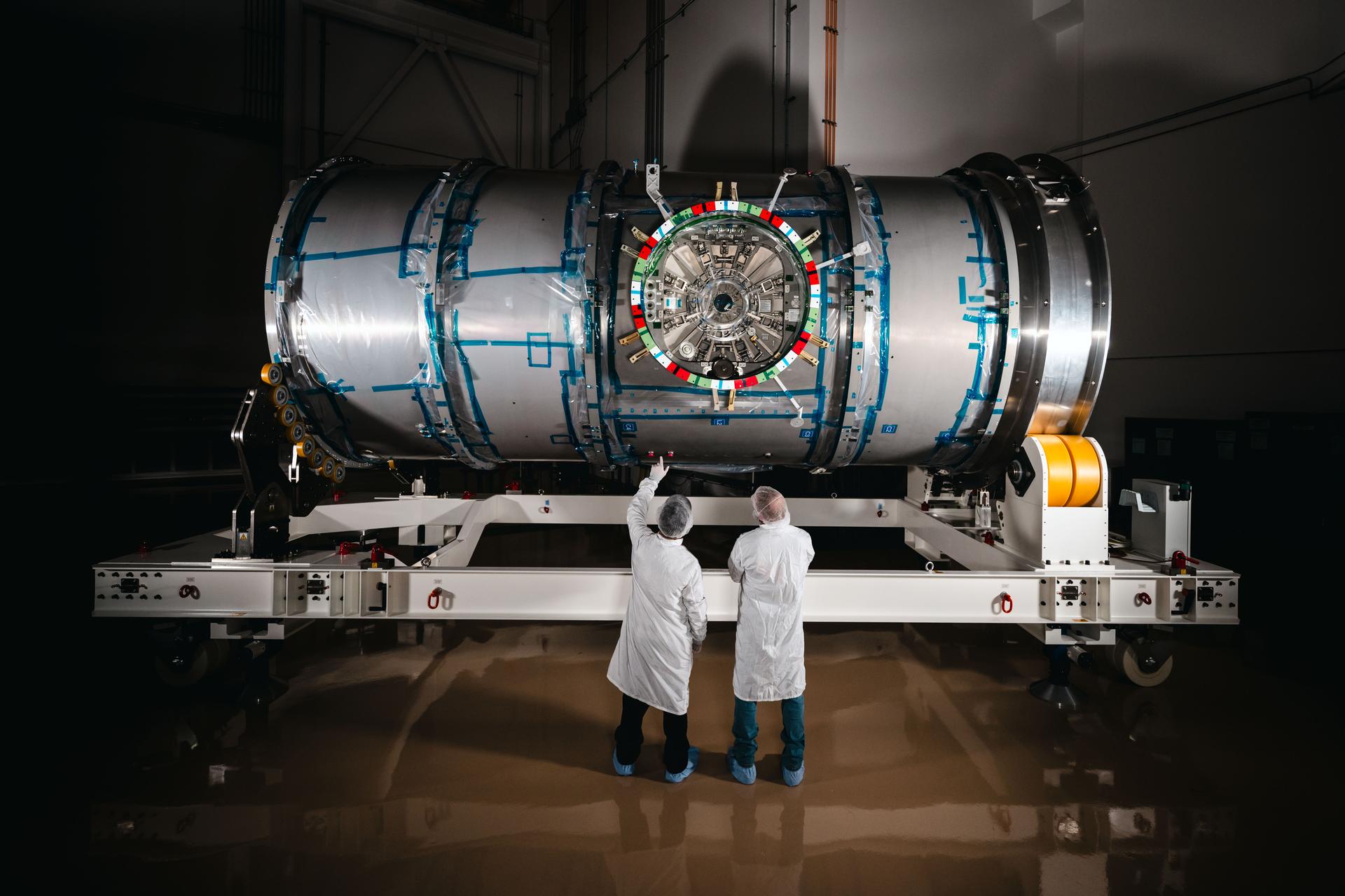 jsc2025e036495large.jpg Two technicians in cleanroom attire stand in front of HALO (Habitation and Logistics Outpost) at Northrop Grumman’s facility in Gilbert, Arizona. The cylindrical metallic module, recently unboxed, rests on a white stand. One technician points toward the front of the module.