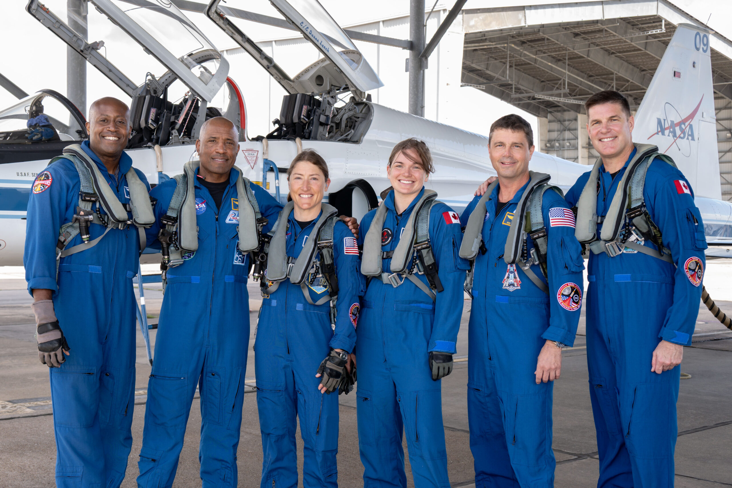 From left to right, NASA astronauts Andre Douglas, Victor Glover, and Christina Koch, CSA (Canadian Space Agency) astronauts Jenni Gibbons, NASA astronaut Reid Wiseman, and CSA astronaut Jeremy Hansen pose in front of an airplane. They are all wearing blue jumpsuits with patches and gray harnesses.
