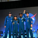 Artemis II Crew Safely Returns to Houston After Historic Mission CSA (Canadian Space Agency) astronaut Jeremy Hansen and NASA astronauts Reid Wiseman, Victor Glover, and Christina Koch smile at the crowd during a news conference. They are all wearing blue jumpsuits with patches on the arms and chest areas. Wiseman raises his right fist in a victorious pose.