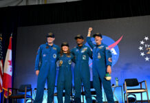 Artemis II Crew Safely Returns to Houston After Historic Mission CSA (Canadian Space Agency) astronaut Jeremy Hansen and NASA astronauts Reid Wiseman, Victor Glover, and Christina Koch smile at the crowd during a news conference. They are all wearing blue jumpsuits with patches on the arms and chest areas. Wiseman raises his right fist in a victorious pose.