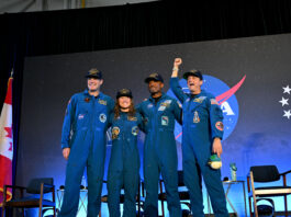 Artemis II Crew Safely Returns to Houston After Historic Mission CSA (Canadian Space Agency) astronaut Jeremy Hansen and NASA astronauts Reid Wiseman, Victor Glover, and Christina Koch smile at the crowd during a news conference. They are all wearing blue jumpsuits with patches on the arms and chest areas. Wiseman raises his right fist in a victorious pose.