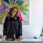 Colombian Judge Pioneers Court Innovation Using Copilot Technology A female judge in robes stands at a desk with a gavel in front of her, behind her a colorful painting and beside her a computer