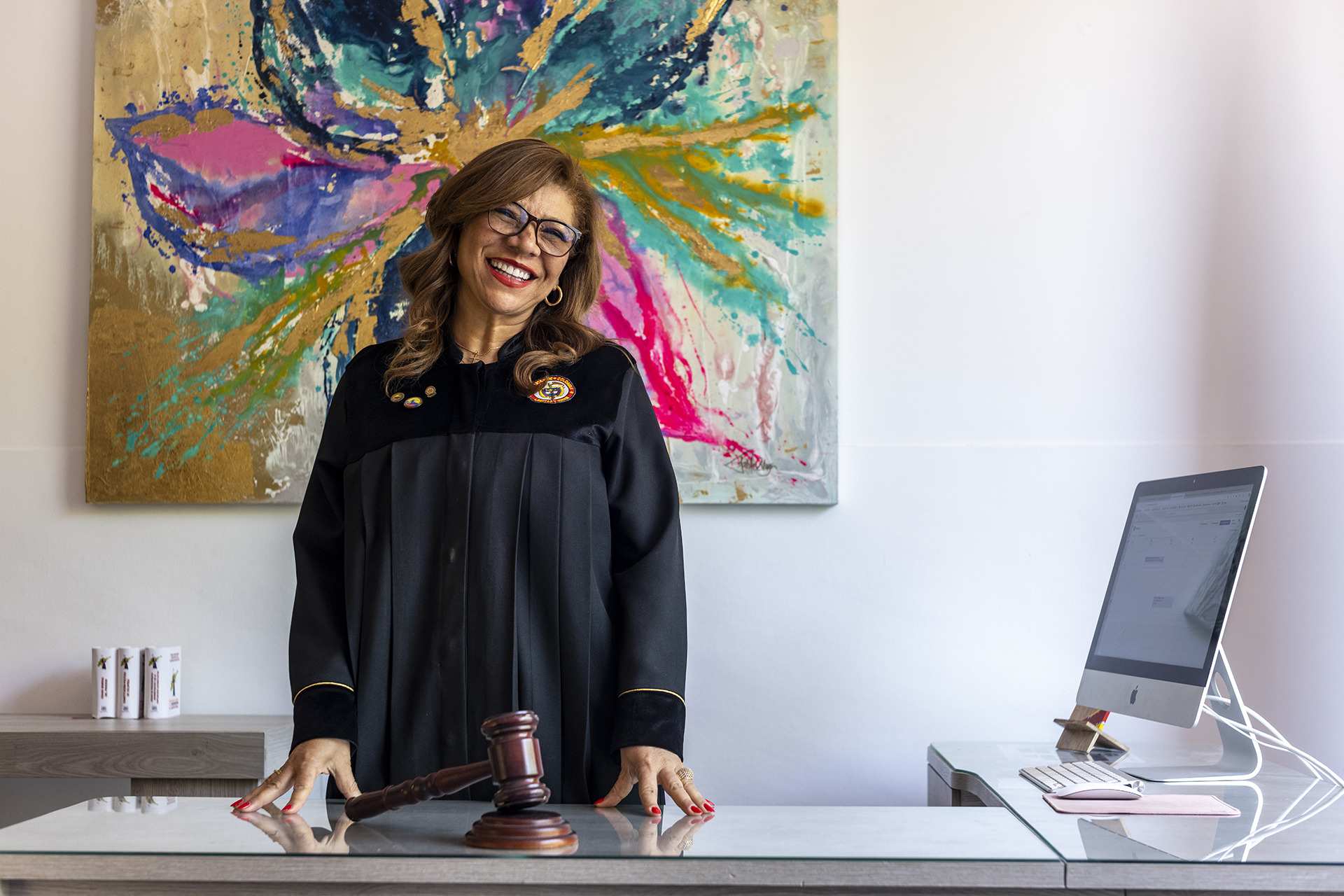 judge maria victoria quinones office 33 1.jpg A female judge in robes stands at a desk with a gavel in front of her, behind her a colorful painting and beside her a computer