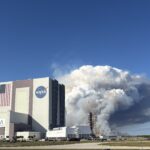 NASA Unveils Advanced Prescribed Burn Technology for Spaceport Operations A large tower of thick white and gray smoke rises behind NASA’s Vehicle Assembly Building at Kennedy Space Center, filling much of the sky. The smoke originates near a launch pad structure with tall cranes beside it. In the foreground, an empty parking lot and several parked vehicles sit under a clear blue sky.