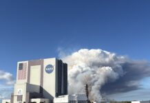 NASA Unveils Advanced Prescribed Burn Technology for Spaceport Operations A large tower of thick white and gray smoke rises behind NASA’s Vehicle Assembly Building at Kennedy Space Center, filling much of the sky. The smoke originates near a launch pad structure with tall cranes beside it. In the foreground, an empty parking lot and several parked vehicles sit under a clear blue sky.