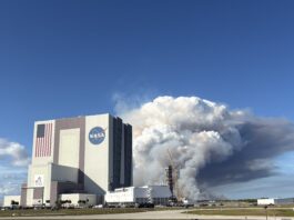 NASA Unveils Advanced Prescribed Burn Technology for Spaceport Operations A large tower of thick white and gray smoke rises behind NASA’s Vehicle Assembly Building at Kennedy Space Center, filling much of the sky. The smoke originates near a launch pad structure with tall cranes beside it. In the foreground, an empty parking lot and several parked vehicles sit under a clear blue sky.