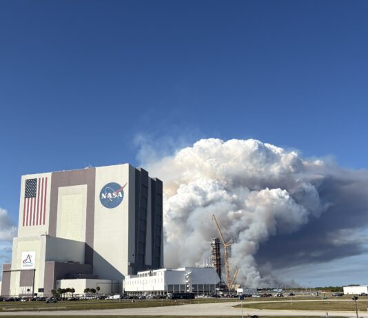 NASA Unveils Advanced Prescribed Burn Technology for Spaceport Operations A large tower of thick white and gray smoke rises behind NASA’s Vehicle Assembly Building at Kennedy Space Center, filling much of the sky. The smoke originates near a launch pad structure with tall cranes beside it. In the foreground, an empty parking lot and several parked vehicles sit under a clear blue sky.