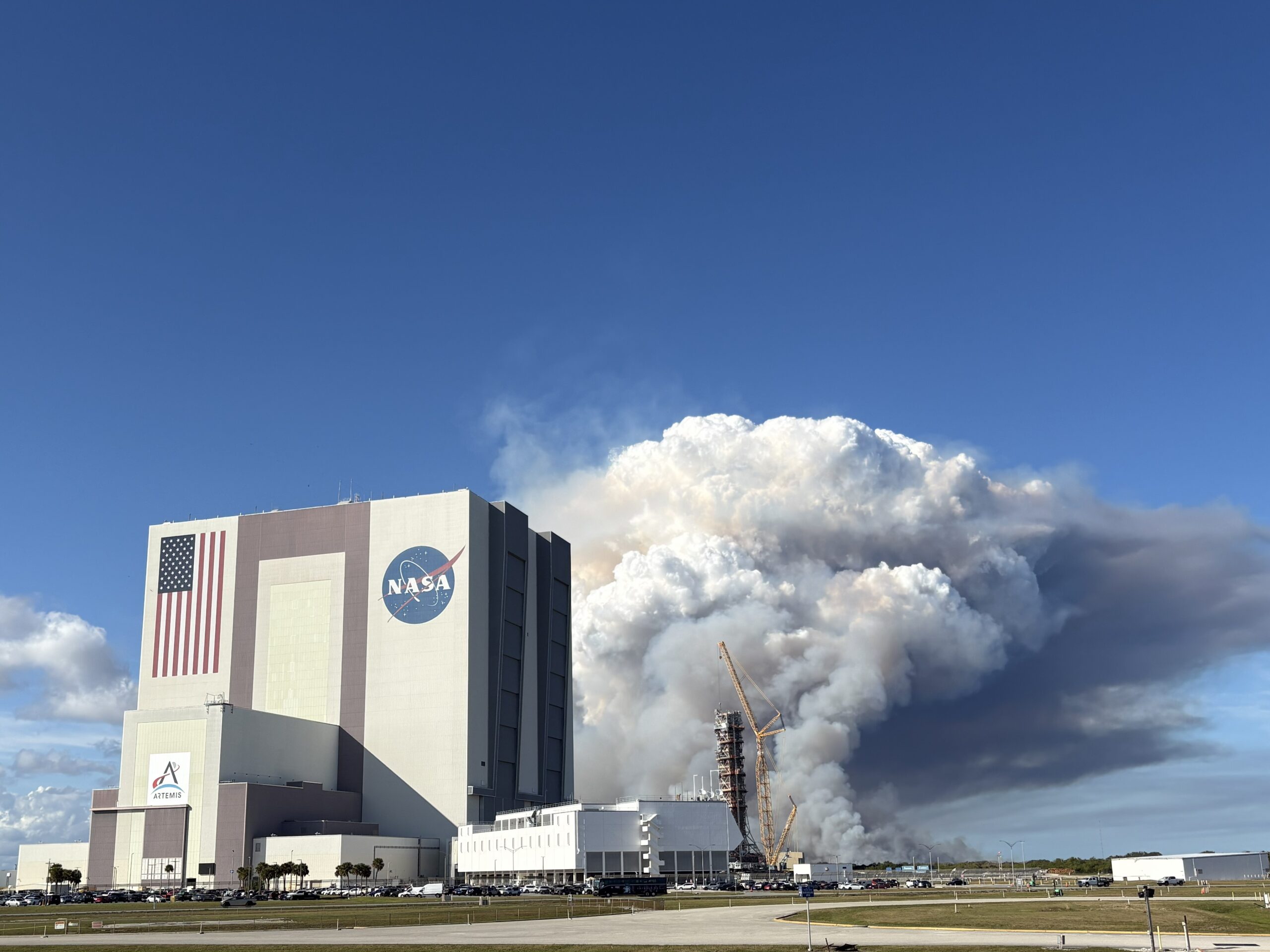 A large tower of thick white and gray smoke rises behind NASA’s Vehicle Assembly Building at Kennedy Space Center, filling much of the sky. The smoke originates near a launch pad structure with tall cranes beside it. In the foreground, an empty parking lot and several parked vehicles sit under a clear blue sky.
